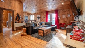 A cozy cabin-style room with a wooden ceiling, a sofa set, a coffee table, and a dining area with a red chair. - Chalet Le Shack Sur la rouge (La Conception)