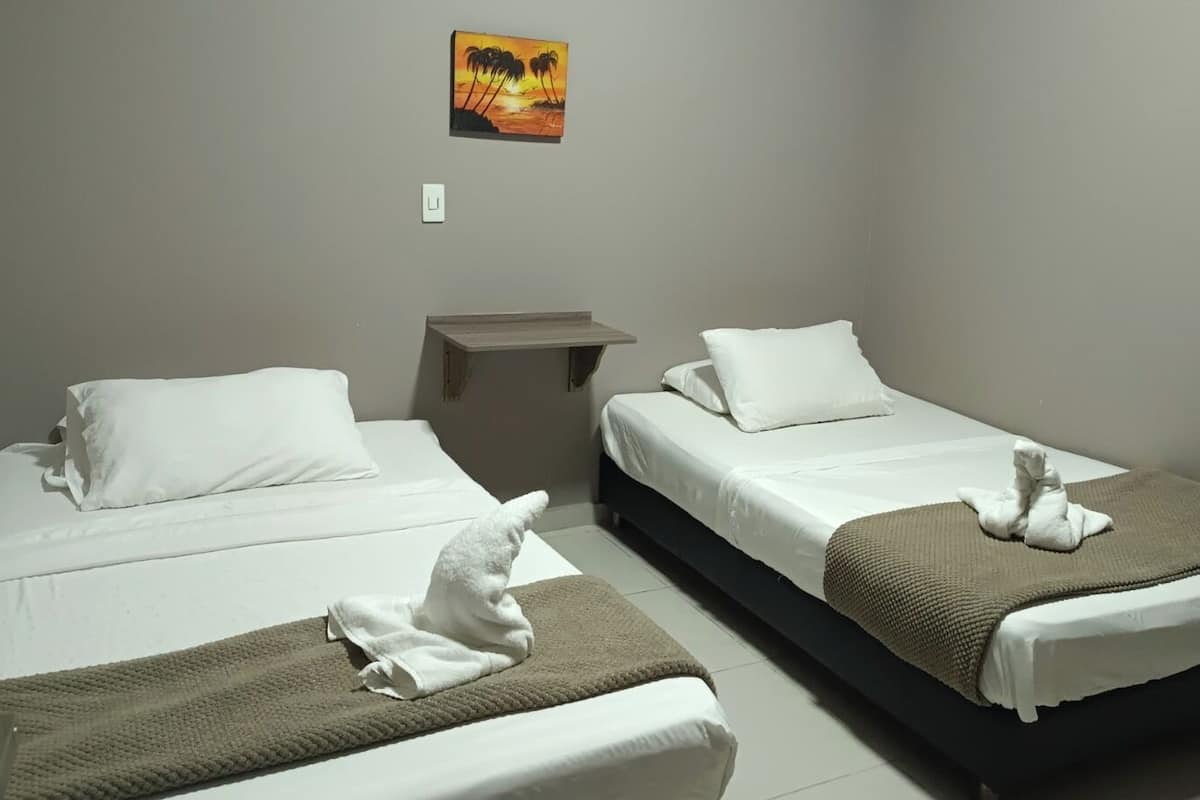 Habitación de hotel con dos camas, cada una con ropa blanca y una manta marrón. Hay una mesita entre las camas y un cuadro en la pared.
