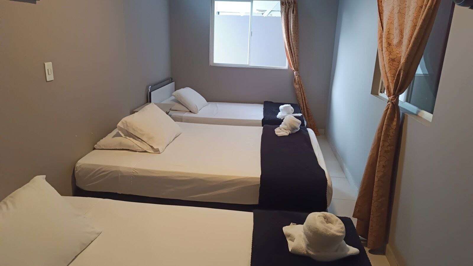 A room with three single beds, each with white bedding and pillows. There is a window with curtains, and a towel is placed on the floor near the beds.