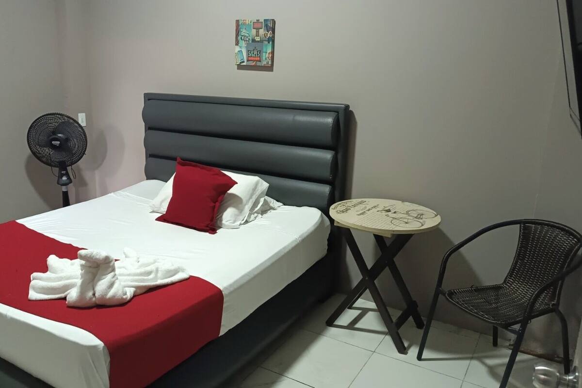 Habitación de hotel con cama, mesita de noche, una silla y un ventilador de pared.