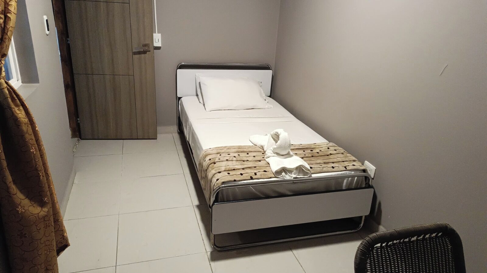 A small, single-bed room with a wooden wardrobe, a white bedspread, and a black chair.