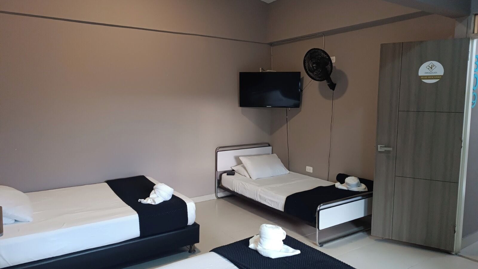 A hotel room with two single beds, a TV mounted on the wall, a wooden wardrobe, and a fan.