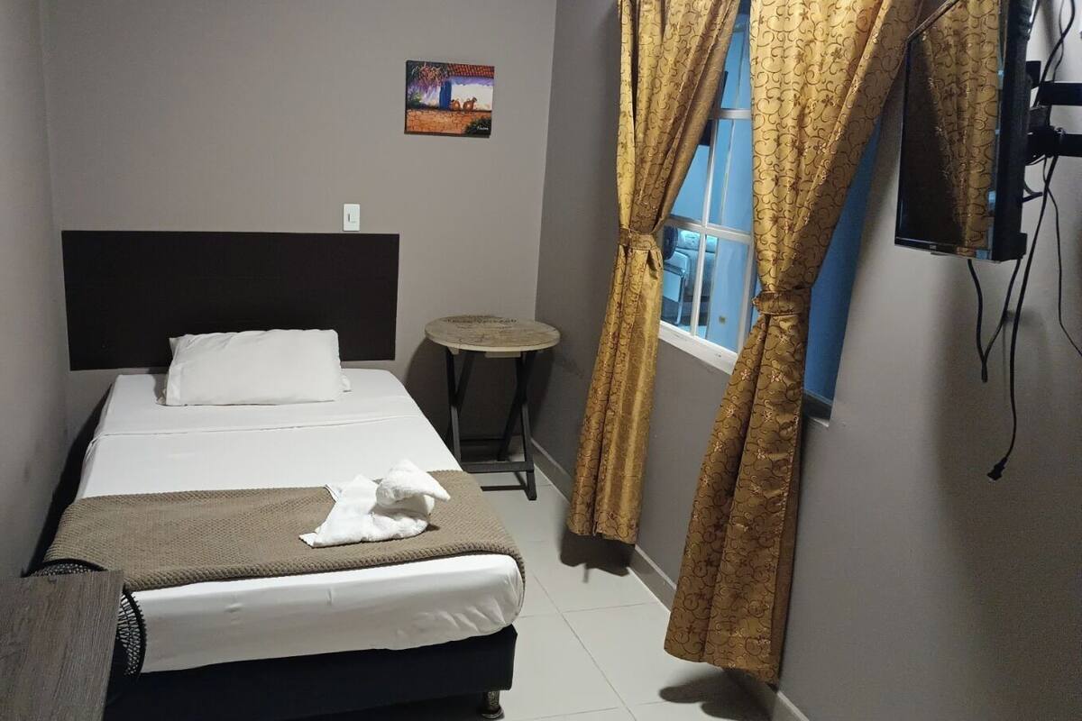 Habitación de hotel con una cama, una mesita redonda pequeña, un televisor y una ventana con cortinas.
