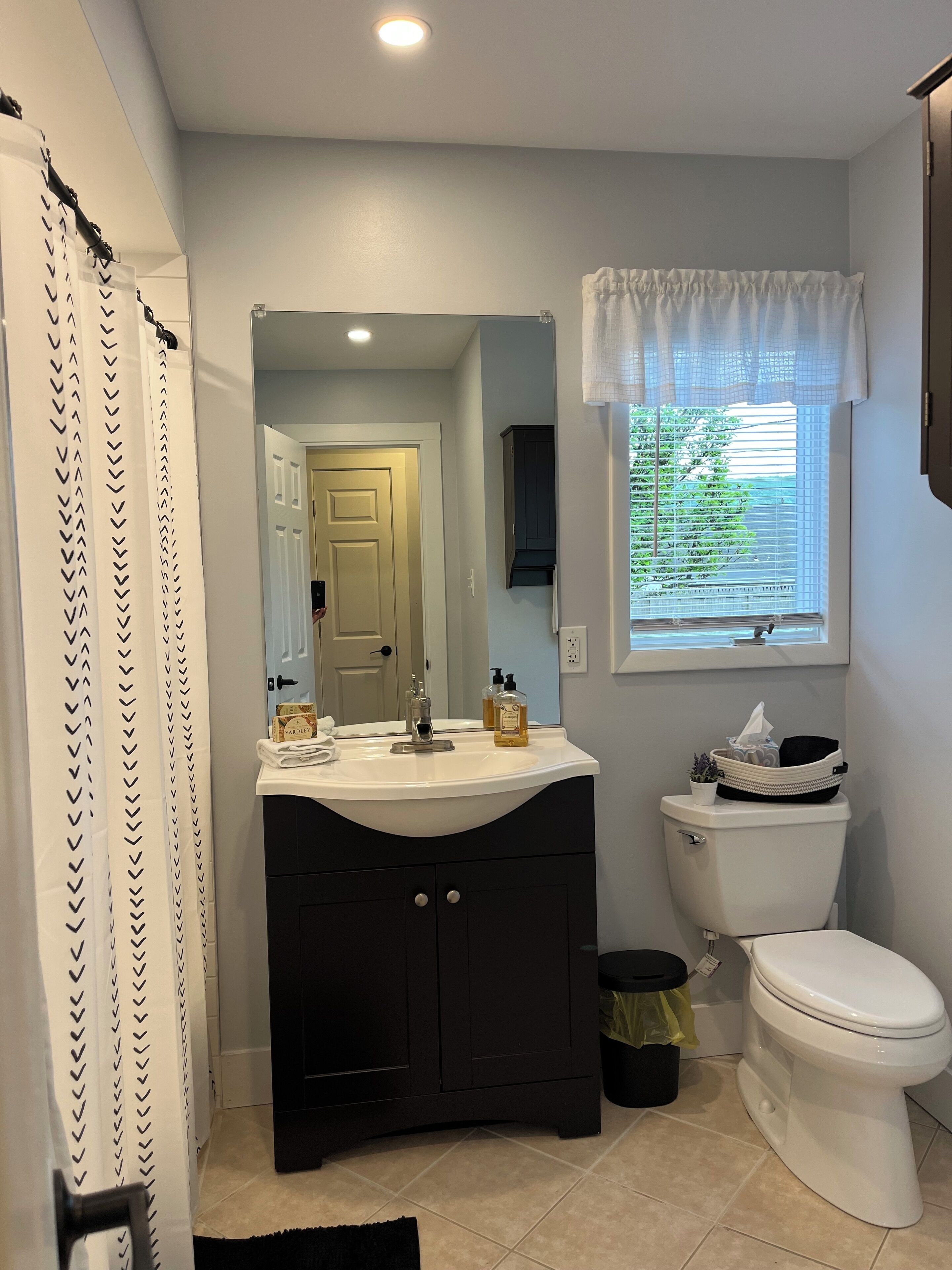 A bathroom with a white sink, dark cabinet, toilet, and a window with a view of greenery.