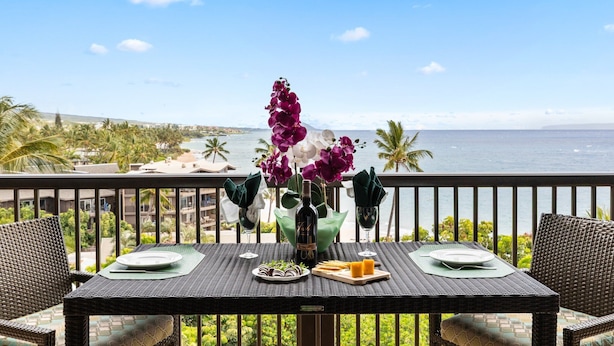 A dining table set with plates, a bottle, and food, overlooking a coastal view with palm trees and a clear sky.