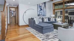 Un salon moderne avec un canapé gris, une table basse et un tapis à motifs. Un escalier avec des rampes en bois mène à un niveau supérieur.