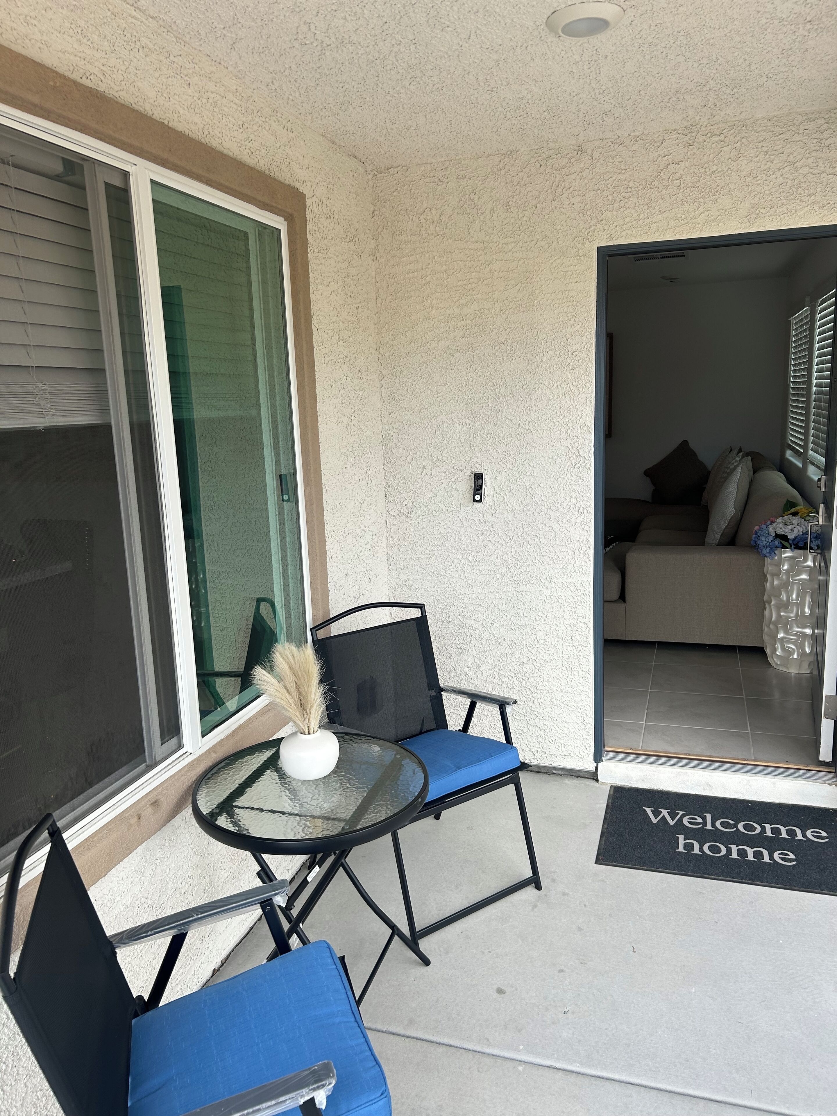 Ein Balkon mit einem Glastisch, zwei Stühlen, einer Fußmatte und Blick ins Wohnzimmer.