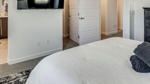 Un dormitorio con cama, ventilador de techo, televisor montado en la pared y puerta que da a otra habitación.