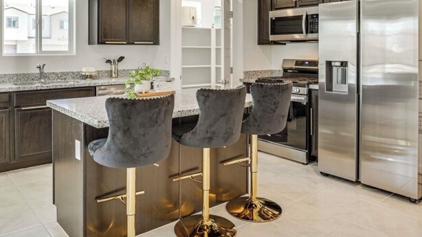 Una cocina moderna con armarios oscuros, un refrigerador de acero inoxidable, un microondas y una encimera de granito. Hay tres taburetes de bar con patas doradas.