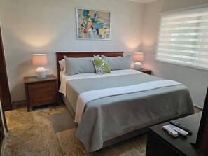 A bedroom with a bed, two bedside tables, a nightstand, a lamp, a painting, and a window with blinds. - Villa Mare B0 on Bavaro Beach, The best tourists location in Bavaro/Punta Cana (Punta Cana)