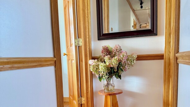 A wooden floor hallway with a door, a small wooden table with a vase of flowers, and a mirror on the wall.