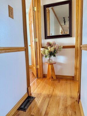 A wooden floor hallway with a door, a small wooden table with a vase of flowers, and a mirror on the wall.