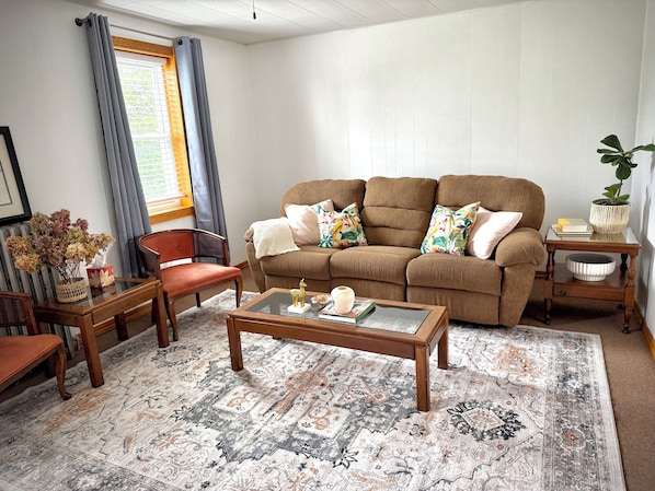 Ein Wohnzimmer mit braunem Sofa, einem Glastisch, einem roten Sessel, einem Holznachttisch und einem gemusterten Teppich.