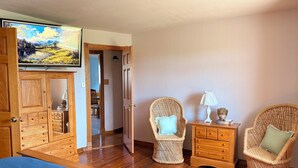 A room with a wooden bed, two wicker chairs, a small wooden nightstand with a lamp, and a painting on the wall.