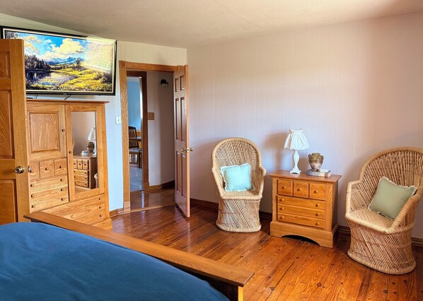 A room with a wooden bed, two wicker chairs, a small wooden nightstand with a lamp, and a painting on the wall.