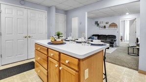 A modern kitchen with a central island, wooden cabinets, and a dining area with a couch and shelves. - Suite Retreat: Cozy Walkable Akron Village Getaway (Akron)