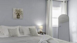 A neatly made bed with white linens, a bedside lamp, a mirror, and a framed floral artwork on the wall. - Suite Retreat: Cozy Walkable Akron Village Getaway (Akron)