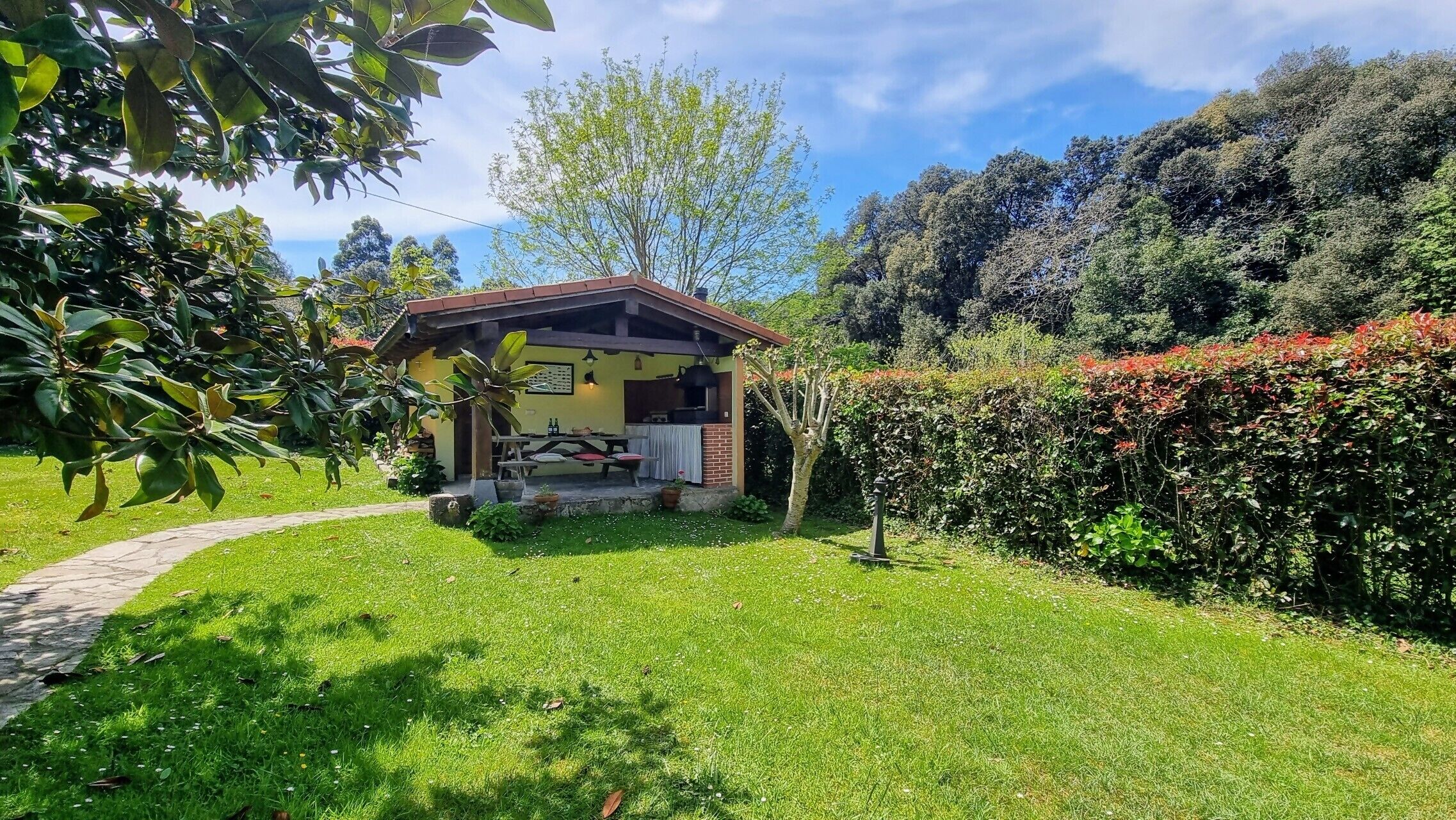 Ein kleines Haus mit Terrasse, umgeben von üppiger Vegetation und blauem Himmel.
