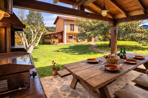 Une terrasse extérieure avec une table en bois dressée pour le repas, un barbecue et une vue sur une maison en arrière-plan.