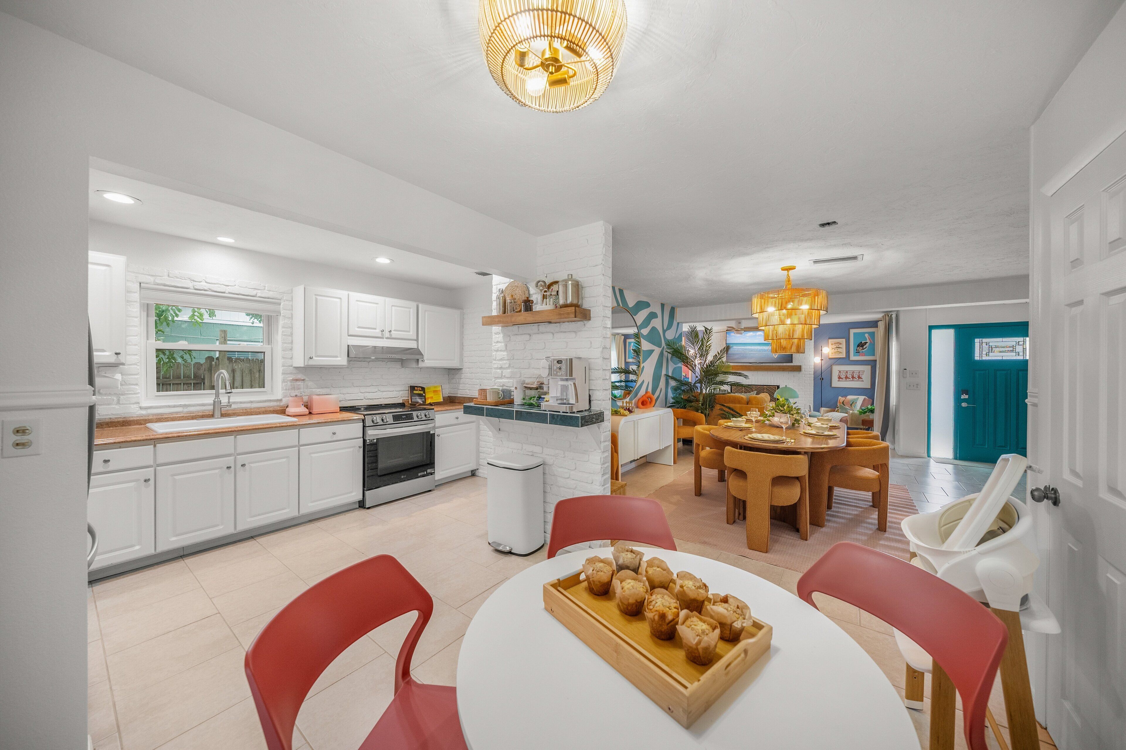Une cuisine moderne avec des meubles blancs, un coin repas avec des chaises rouges et un îlot central sur lequel se trouve un plateau de muffins.