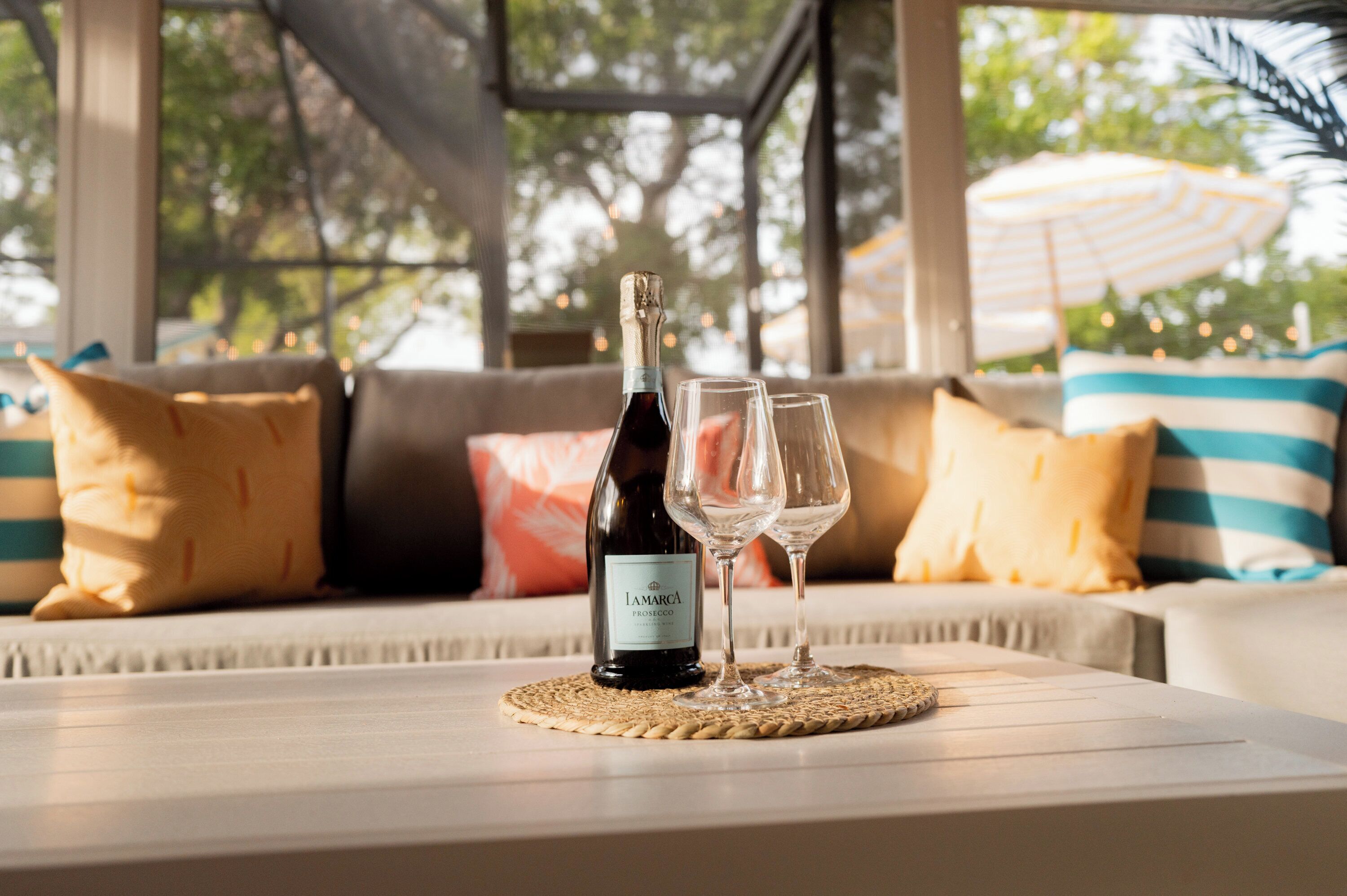 Une bouteille de vin et deux verres sur une table, dans une pièce chaleureuse et lumineuse, avec vue sur les arbres et un parasol à l’extérieur.