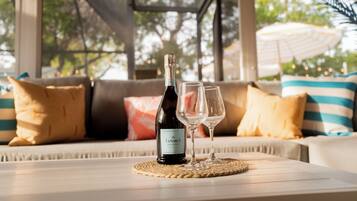 Une bouteille de vin et deux verres sur une table, dans une pièce chaleureuse et lumineuse, avec vue sur les arbres et un parasol à l’extérieur.