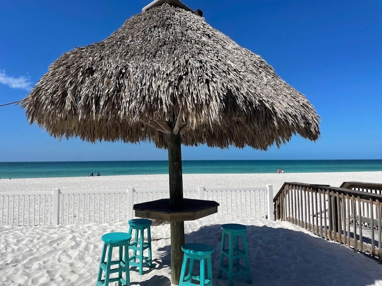 Ein Strandkorb, ein Holztisch und zwei türkisfarbene Hocker am sandigen Strand mit einer Holzlattenveranda und blauem Himmel.