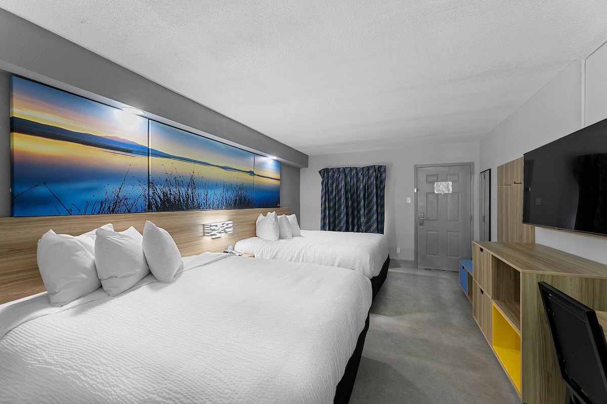 A hotel room with two beds, a large wall art of a sunset, a TV, and a wooden desk.