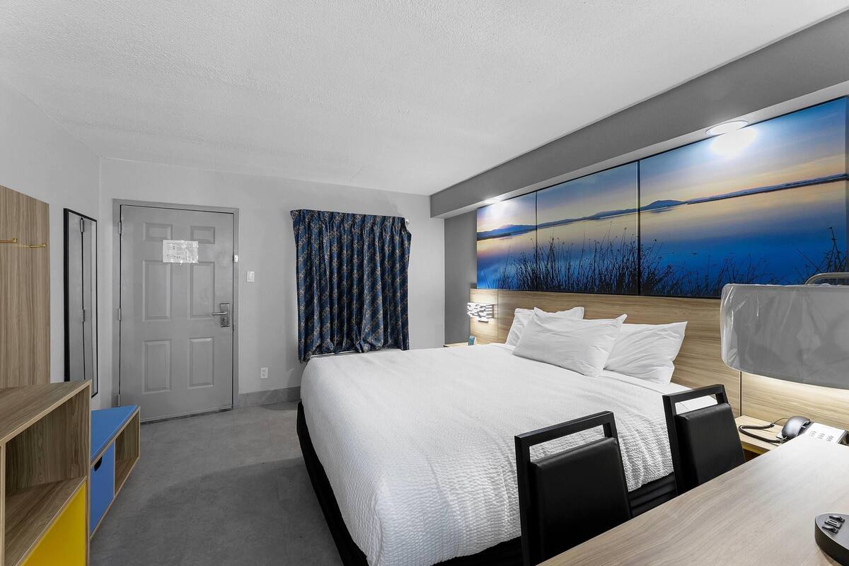 A hotel room with a large bed, a desk with a computer, a desk lamp, and a painting of a sunset on the wall.