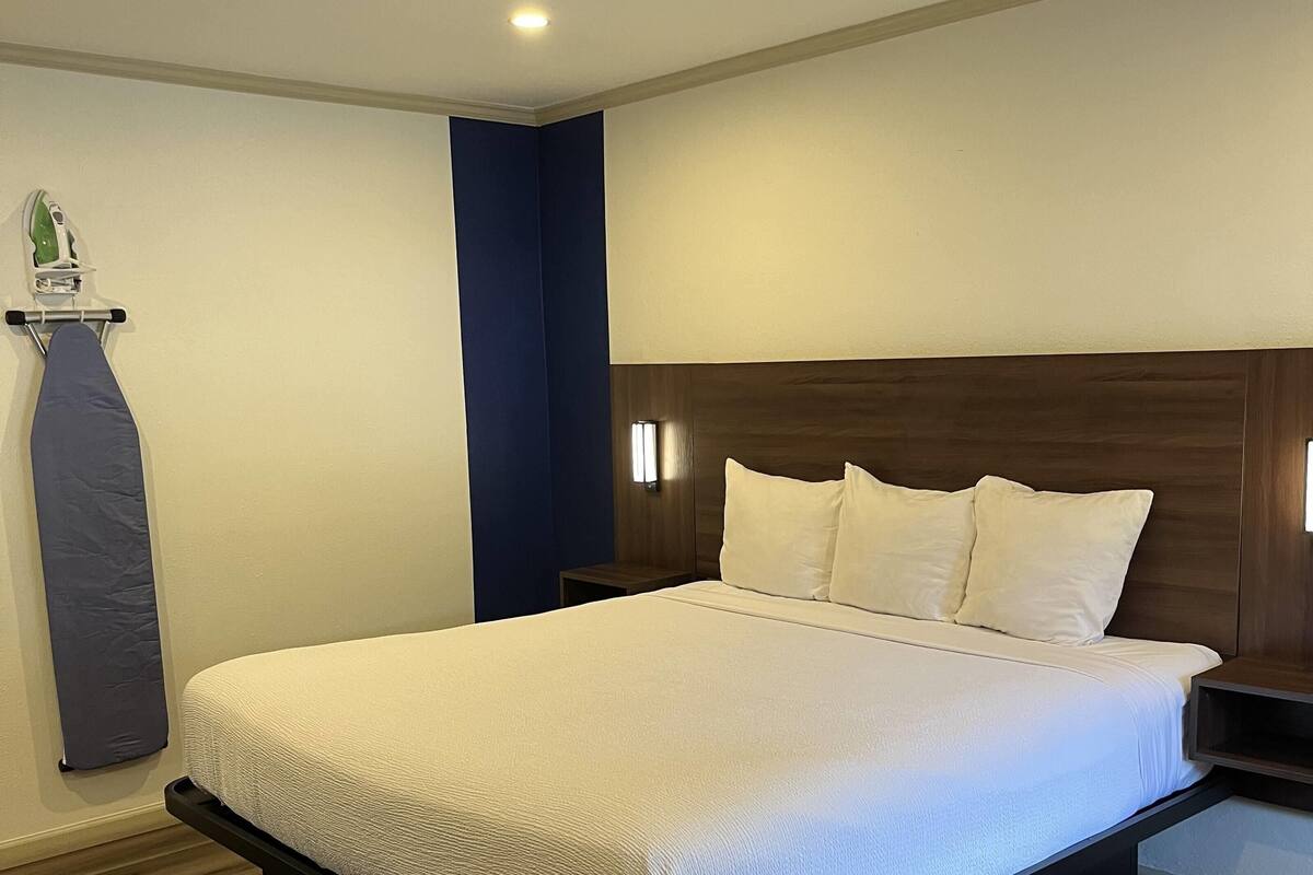 Una cama bien hecha con sábanas blancas, un cabecero de madera y una mesita de noche con una lámpara.