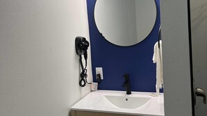 A bathroom with a white sink, a black faucet, a round mirror, and a hairdryer on a wall-mounted charging station. - Travelodge By Wyndham Bainbridge (Bainbridge)