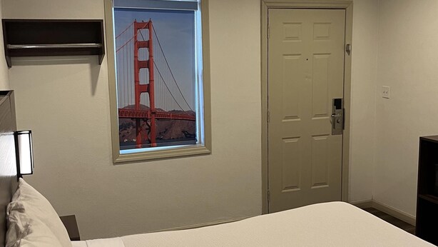A bedroom with a bed, a door, a wall-mounted shelf, and a window with a view of the Golden Gate Bridge. - Travelodge By Wyndham Bainbridge (Bainbridge)