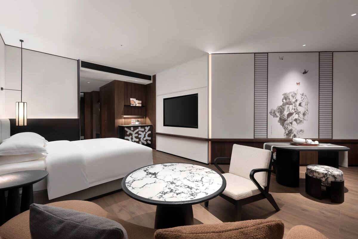 Quarto de hotel moderno com uma cama grande, uma mesa redonda de mármore, um sofá e uma televisão na parede.
