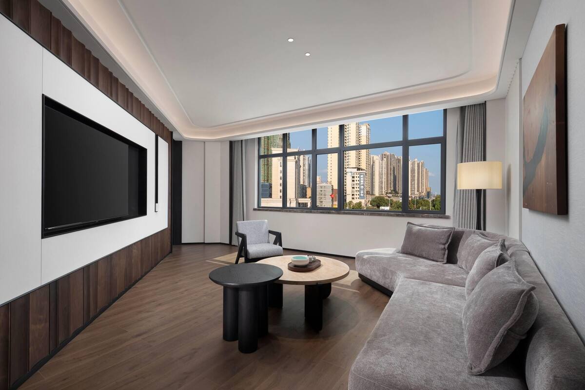 Uma sala de estar moderna com uma televisão grande, assentos confortáveis, uma mesa de centro redonda e vista para edifícios altos através de grandes janelas.