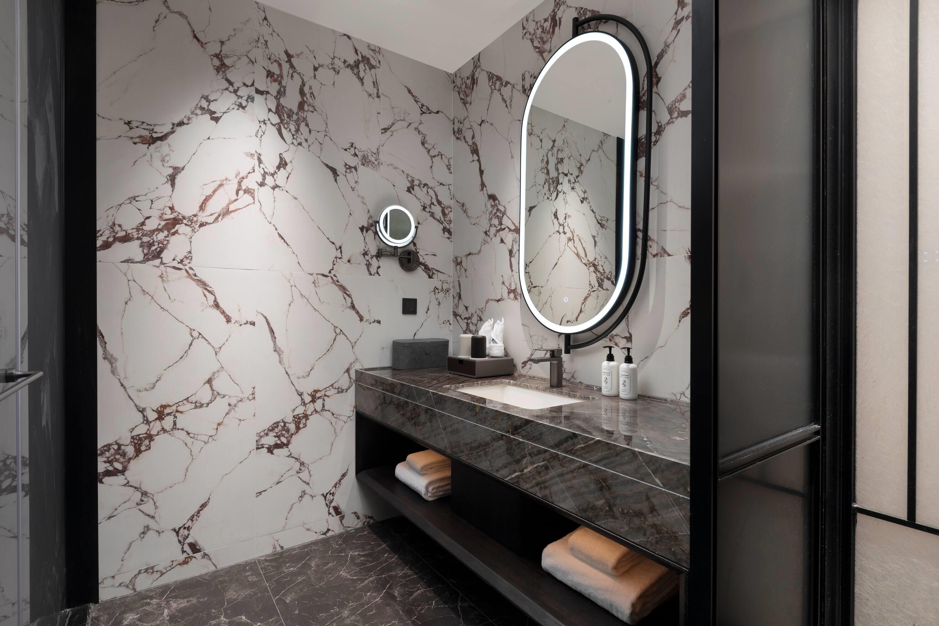 A modern bathroom with a marble wall, a large oval mirror, a black vanity, and a towel rack.