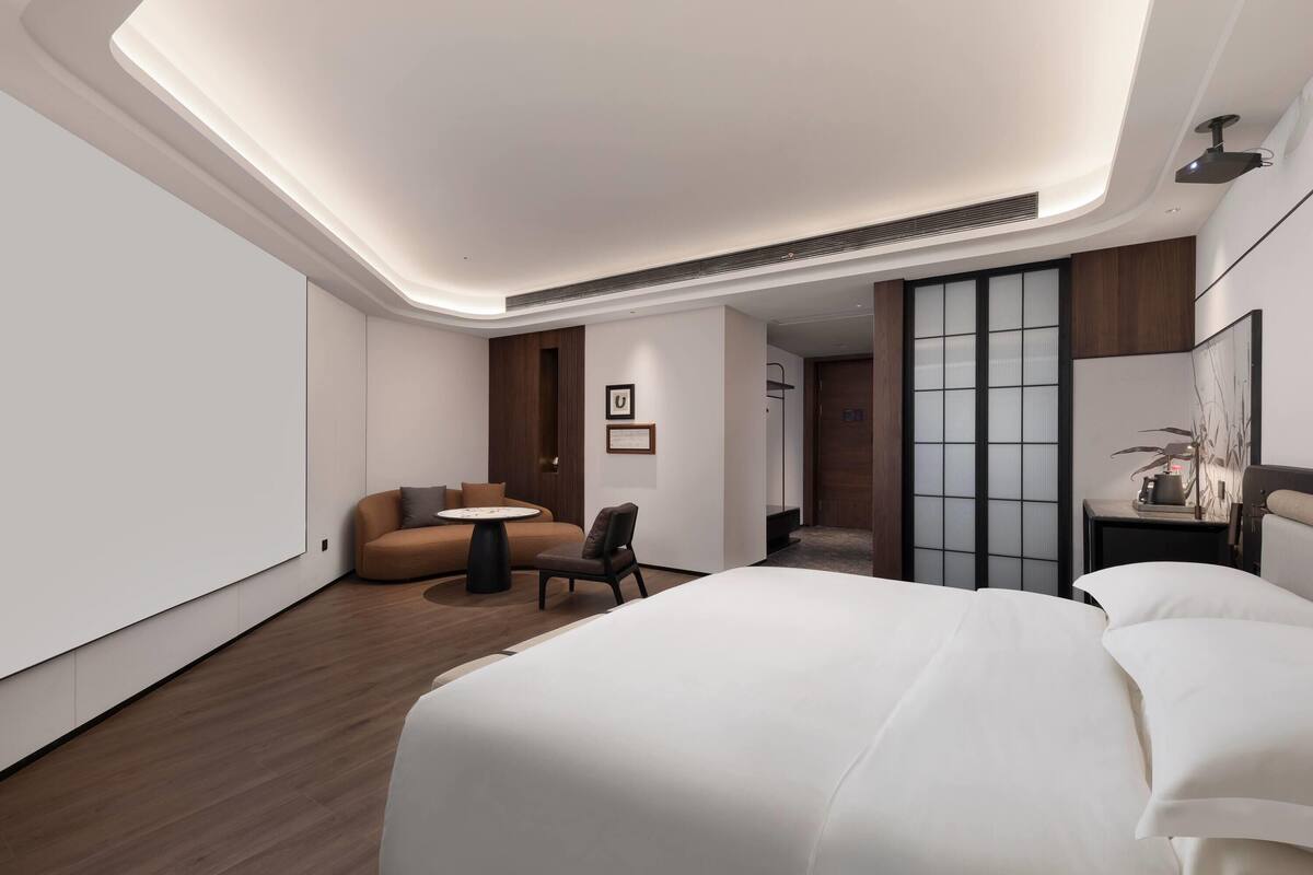 Quarto de hotel moderno com uma cama grande, um sofá, uma mesinha e uma cadeira. Há uma porta deslizante que dá para outro quarto.