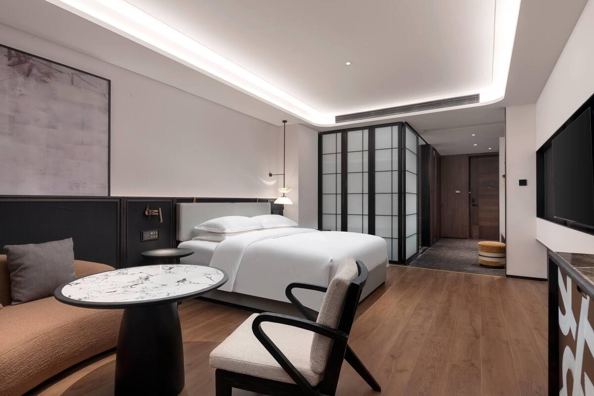 Quarto de hotel moderno com cama, sofá, mesa redonda e televisão.