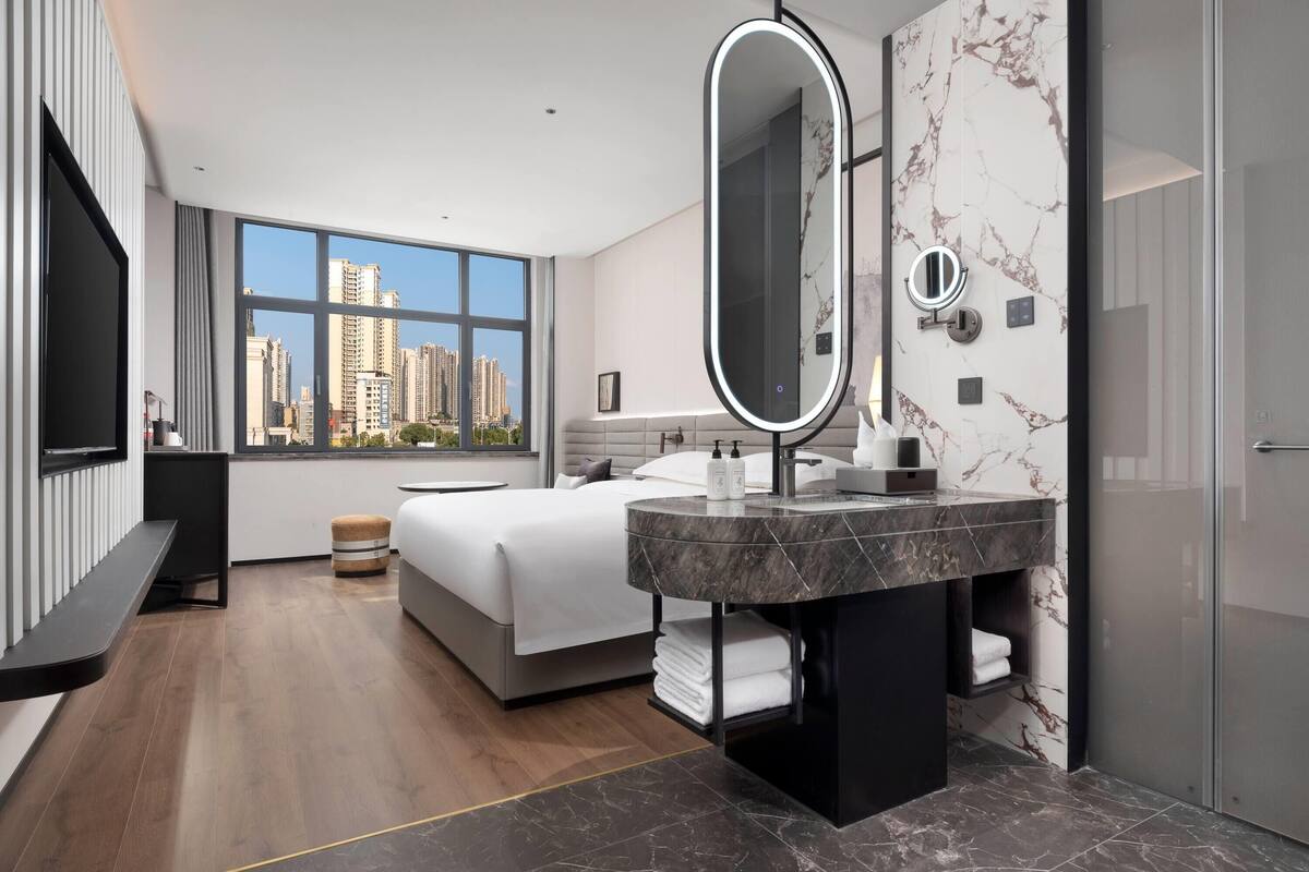 Quarto de hotel moderno com uma cama grande, um lavatório de mármore redondo, um espelho grande e vista para edifícios altos pela janela.