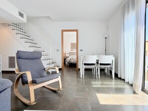 A modern living room with a wooden rocking chair, a dining table with chairs, a staircase, and a view through a door. - Roca Maura by Costabravaway AC /Sea view (L'estartit)