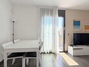 Una habitación moderna y bien iluminada con una mesa de comedor, sillas, un televisor de pantalla plana y un balcón con cortinas.