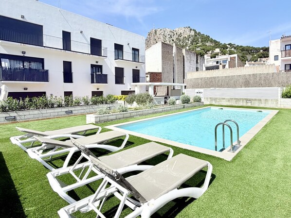 Una piscina con sillas de descanso, un edificio con balcones y una montaña al fondo.