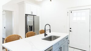 A modern kitchen with white cabinetry, a stainless steel refrigerator, a sink, and wicker chairs.