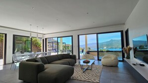 A modern living room with a large sectional sofa, a coffee table, and a flat-screen TV. The room has large windows offering a view of the outdoors. - Villa Haut Standing Stella Di Mare panoramic view (Propriano)