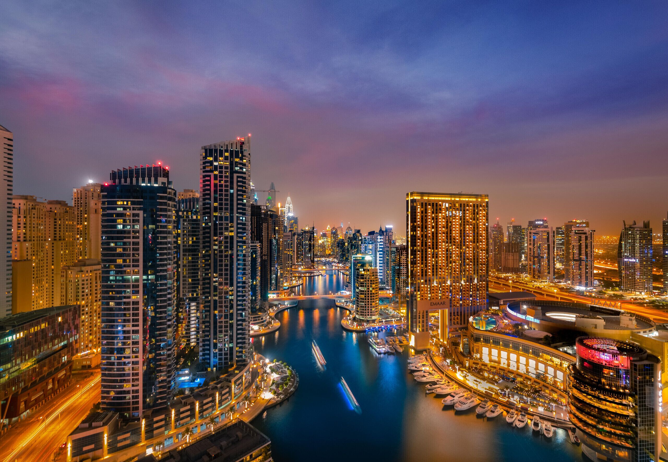 A cityscape at dusk with high-rise buildings, a marina with boats, and a river.
