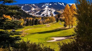 A golf course with a mountain backdrop, surrounded by autumn trees and a clear sky.