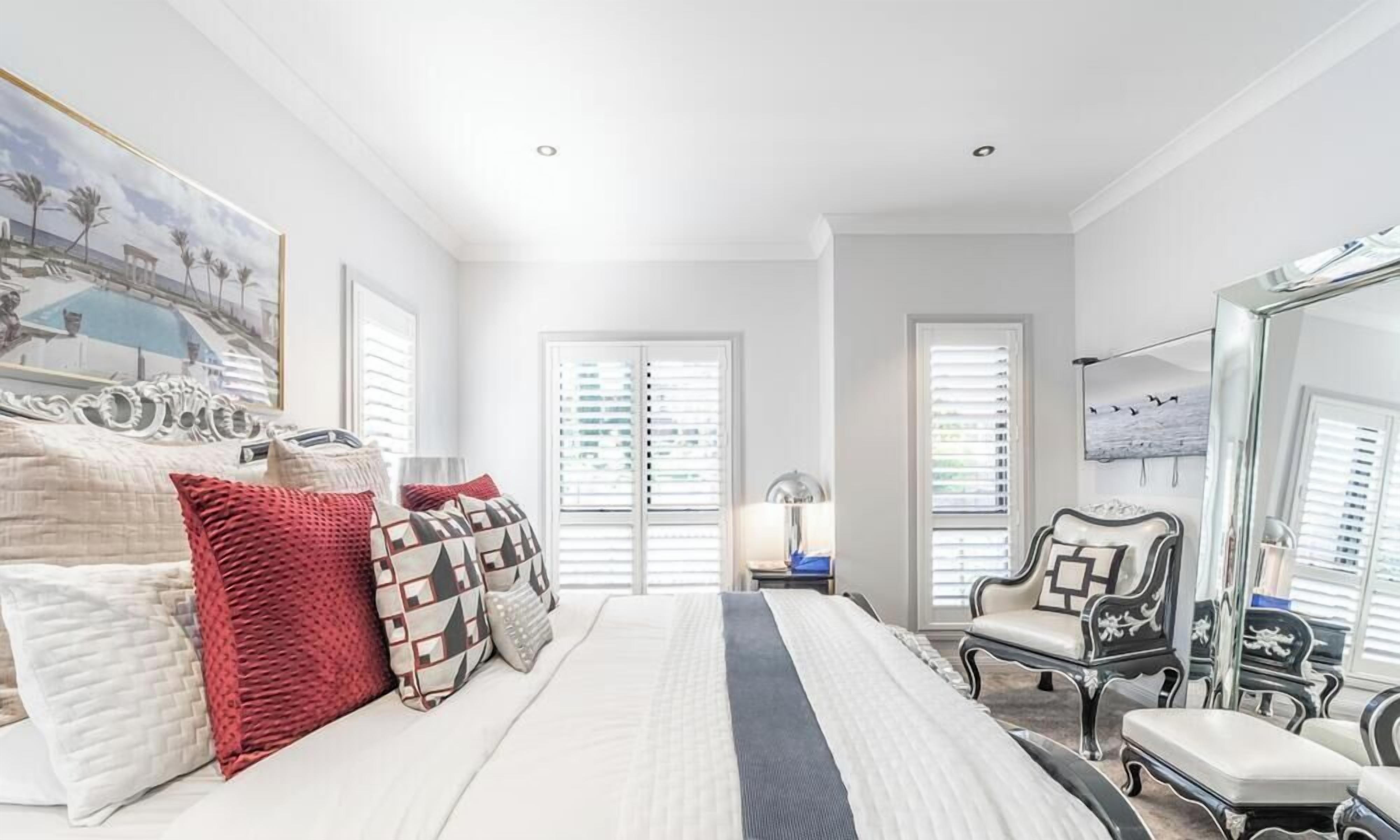 A bedroom with a large bed, two windows with blinds, a chair, and a mirror.