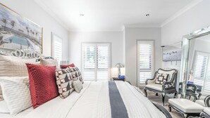 Un dormitorio con una cama grande, dos ventanas con persianas, una silla y un espejo.