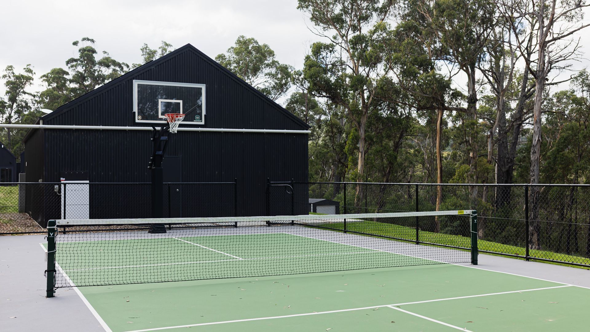 Un campo da tennis all'aperto con una rete, un canestro da basket e un edificio nero sullo sfondo.