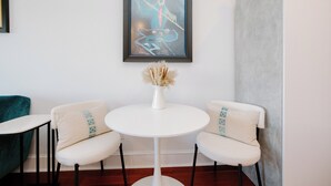A room with a white round table, two white cushioned chairs, a vase with dried flowers, and a framed artwork on the wall.