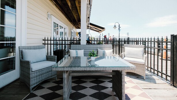 A covered outdoor seating area with wicker furniture and a glass table, adjacent to a building with large windows and a checkered floor.